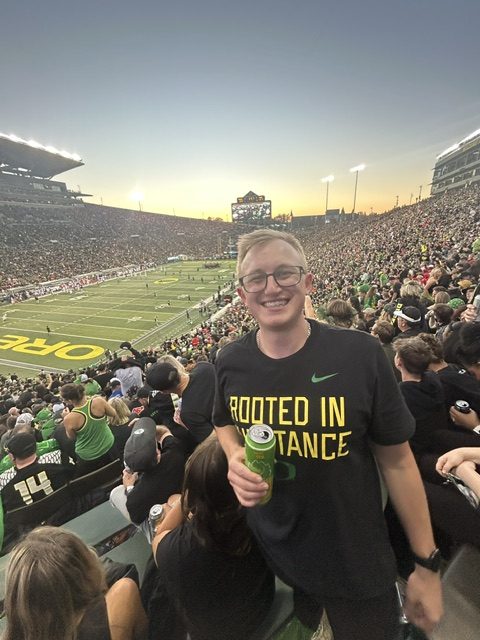 Hayden Schuster smiling in the stands at Autzen Stadium at sunset, holding a drink