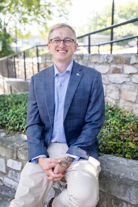 Hayden Schuster smiling, seated on a stone wall in a blue blazer