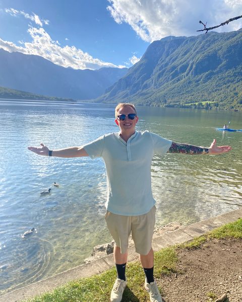 Hayden Schuster with his arms spread wide at the edge of a clear mountain lake