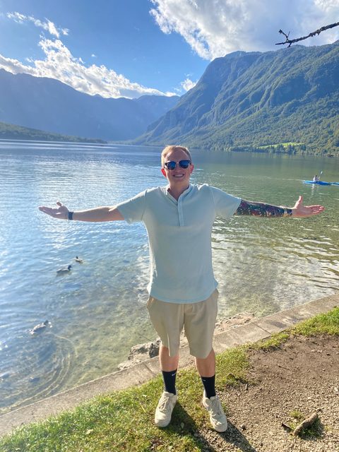 Hayden Schuster with his arms spread wide at the edge of a clear mountain lake